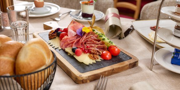 A beautifully arranged plate with various types of cold cuts, tomatoes, and grapes. In the background, rolls and additional dishes can be seen on a table. - Wirtshaus am Giggijoch - Sölden