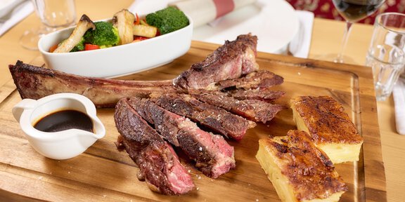 A delicious meat dish on a wooden board, accompanied by a sauce and a side of vegetables and bread. The table is elegantly set with glasses and cutlery. - Wirtshaus am Giggijoch - Sölden