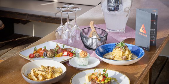 A stylishly set table with various dishes, including pasta, meat, and salad. In the background, there are glasses and a cool bottle of water. - Wirtshaus am Giggijoch - Sölden