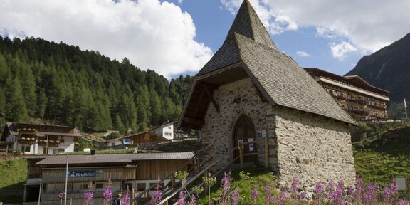 An old stone church with a pointed roof stands in front of a picturesque mountain landscape. Colorful flowers bloom in the foreground, and traditional houses can be seen in the background. - Bergsteigerkapelle - Ausstellung - Vent