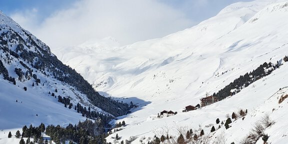 Eine verschneite Berglandschaft mit hohen Gipfeln und bewaldeten Hängen. Im Tal sind einige Gebäude sichtbar, umgeben von Schnee. - Kunstweg Barteb'ne - Vent