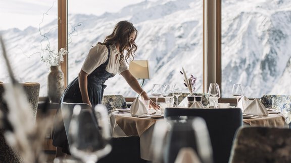 Eine Kellnerin deckt einen Tisch in einem Restaurant mit Blick auf schneebedeckte Berge. Die Atmosphäre ist gemütlich und einladend. - SKI | GOLF | WELLNESS Hotel Riml - Hochgurgl