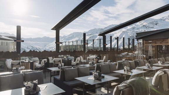Eine moderne Terrasse eines Restaurants mit Blick auf schneebedeckte Berge. Die Tische sind elegant gedeckt und die Sonne scheint auf die gemütlichen Sitzgelegenheiten. - SKI | GOLF | WELLNESS Hotel Riml - Hochgurgl