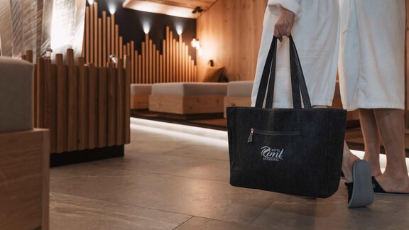 Eine Person in einem Bademantel trägt eine große Tasche in einem modernen, eleganten Spa-Bereich. Der Raum ist mit Holz und sanftem Licht gemütlich gestaltet. - SKI | GOLF | WELLNESS Hotel Riml - Hochgurgl