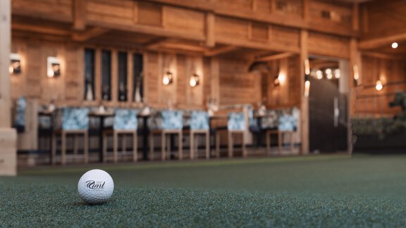 Ein Golfball liegt auf einem grünen Plätzchen im Vordergrund. Im Hintergrund sind eine rustikale Einrichtung und Tische in einem gemütlichen Raum zu sehen. - SKI | GOLF | WELLNESS Hotel Riml - Hochgurgl
