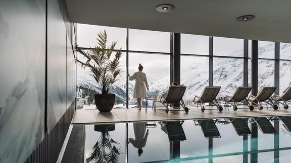 Ein eleganter Spa-Bereich mit einem Pool und großen Fenstern, die auf schneebedeckte Berge blicken. Eine Person in einem Bademantel steht entspannt und genießt die Aussicht. - SKI | GOLF | WELLNESS Hotel Riml - Hochgurgl