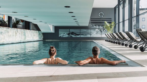 Ein moderner Innenpool mit entspannenden Liegen und großen Fenstern. Zwei Personen genießen die Ruhe am Beckenrand. - SKI | GOLF | WELLNESS Hotel Riml - Hochgurgl