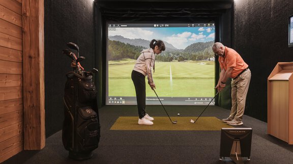 Eine Innen-Golfanlage mit einem Simulator. Ein Mann und eine Frau üben das Golfspielen auf der virtuellen Plattform. - SKI | GOLF | WELLNESS Hotel Riml - Hochgurgl