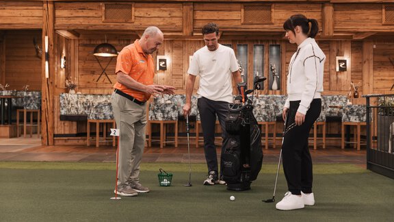 Ein Golftrainer zeigt zwei Personen, wie man puttet. Sie befinden sich in einer holzverkleideten Indoor-Golfumgebung. - SKI | GOLF | WELLNESS Hotel Riml - Hochgurgl