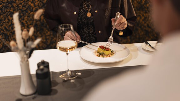 Eine Person sitzt an einem Tisch mit einem eleganten Gericht. Sie hält Besteck und genießt ein Glas Wein. - SKI | GOLF | WELLNESS Hotel Riml - Hochgurgl