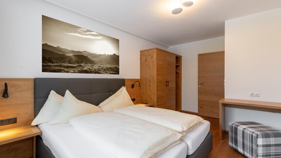 Ein modernes Schlafzimmer mit einem großen Doppelbett und Holzdetails. Eine Fotografie von Bergen hängt an der Wand, und es gibt einen Schrank sowie einen kleinen Hocker im Raum. - Alpenappartement Central Valley Penthouse - Längenfeld