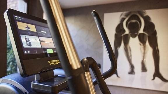 A modern fitness device with a screen that stands in a stylish room. In the background, there is an artistic image of a sprinter. - Art & Relax Hotel Bergwelt - Obergurgl 