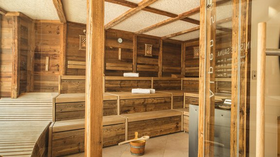 A cozy sauna with wood paneling and several benches. In the foreground are a bucket and cushions for relaxing. - Art & Relax Hotel Bergwelt - Obergurgl 