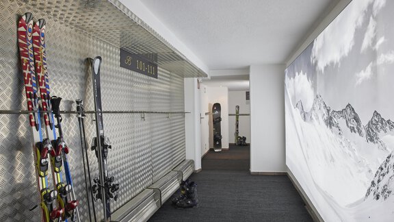A modern ski storage room with skis and snowboards on the wall. In the background, there is a large picture of snow-covered mountains. - Art & Relax Hotel Bergwelt - Obergurgl 