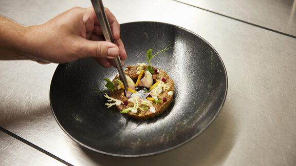 An upscale dish in a black bowl is garnished with tweezers. The food contains fresh herbs and colorful ingredients. - Art & Relax Hotel Bergwelt - Obergurgl 