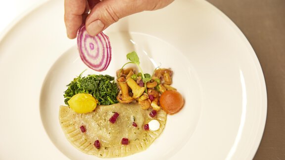 An artistic dish on a white plate with a handcrafted ravioli, vegetables, and a fresh garnish. A person adds a slice of red onion. - Art & Relax Hotel Bergwelt - Obergurgl 