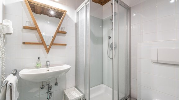 Ein modernes Badezimmer mit einer Duschkabine und einem Waschbecken. Die Wände sind weiß gefliest und es gibt ein stilvolles, dreieckiges Spiegel-Design. - Ferienwohnung Alt Poschach - Obergurgl 