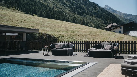 A modern outdoor area with a pool and comfortable lounge furniture. In the background, green hills and a blue sky can be seen. - The Crystal VAYA Unique - Obergurgl 