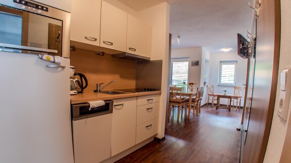 A modern kitchen with white cabinets and wooden flooring. In the background, a dining area with a table and chairs is visible. - Alpenapart Saphir - Sölden