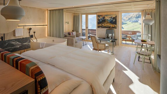 A modern hotel room with a large bed and cozy seating area. Large windows offer a view of the mountains and the outdoor area. - Alpengasthof Grüner - Sölden