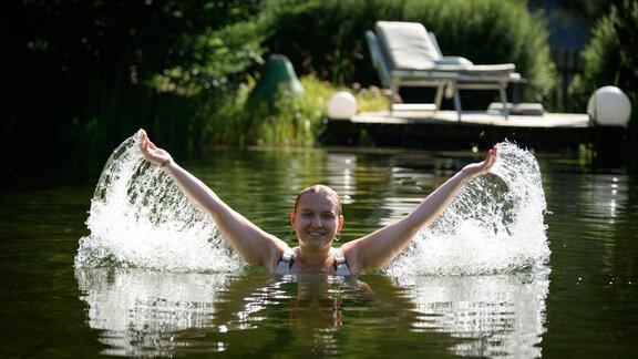 A woman is swimming happily in the clear water and splashing with her arms. In the background, there are lounge chairs and green plants visible. - Alpengasthof Grüner - Sölden