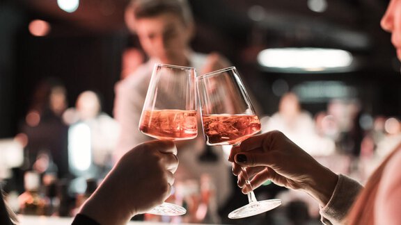Zwei Gläser mit roséfarbener Flüssigkeit stoßen an. Im Hintergrund sind unscharfe Silhouetten von Menschen zu sehen. - AlpenParks The Secret Sölden - Sölden