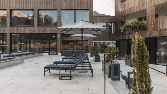 Eine moderne Terrasse mit Sonnenliegen und Sonnenschirmen. Im Hintergrund sind Gebäude mit Holzfassaden zu sehen. - AlpenParks The Secret Sölden - Sölden