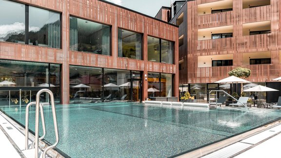 Ein eleganter Außenpool umgeben von modernen Gebäuden. Sonnenschirme und Liegen laden zum Entspannen ein. - AlpenParks The Secret Sölden - Sölden