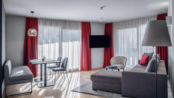 Ein modernes Wohnzimmer mit einer gemütlichen Couch und einem Esstisch. Große Fenster lassen viel Licht herein und die roten Vorhänge sorgen für einen auffälligen Akzent. - AlpenParks The Secret Sölden - Sölden