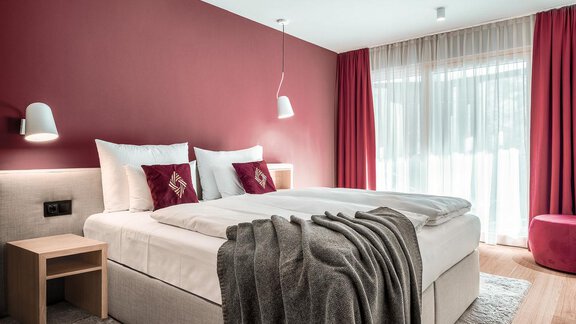 Ein modernes Schlafzimmer mit einem gemütlichen Doppelbett und roten Akzenten. Die Fenster sind mit hellen Vorhängen ausgestattet, die viel Licht hereinlassen. - AlpenParks The Secret Sölden - Sölden