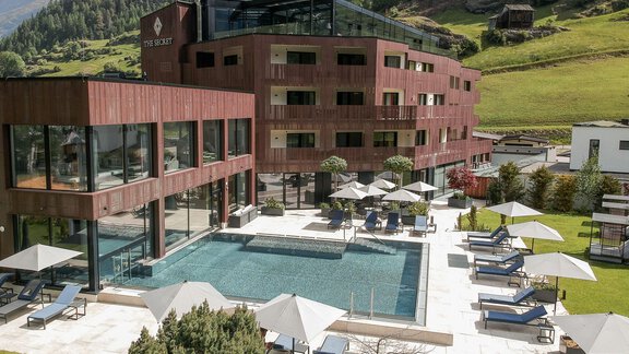Ein modernes Hotel mit einem großen Schwimmbad und Liegen im Garten. Im Hintergrund sind grüne Berge zu sehen. - AlpenParks The Secret Sölden - Sölden
