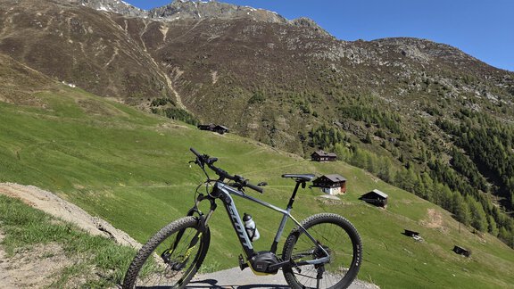 Ein Mountainbike steht auf einem Weg in einer grünen Berglandschaft. Im Hintergrund sind die Berge und einige Hütten zu sehen. - Apart Alpenjuwel - Sölden