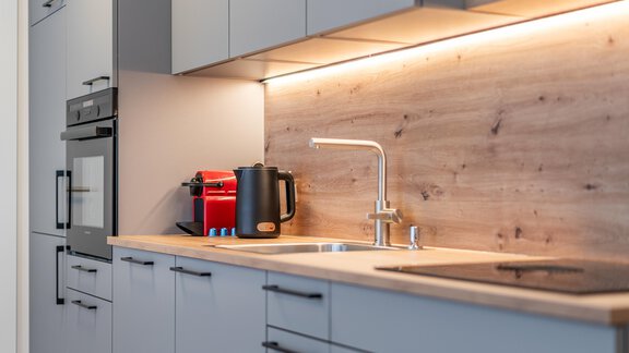 Eine moderne Küche mit grauen Schränken und Holzdetails. Auf der Arbeitsplatte stehen ein Wasserkocher und eine Kaffeemaschine. - Apart Sonnenbichl - Sölden