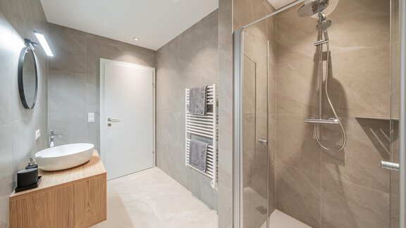 Ein modernes Badezimmer mit einer begehbaren Dusche und einem Holzwaschbecken. Die Wände sind in einem neutralen Grau gehalten und das Licht ist hell und einladend. - Apart Sonnenbichl - Sölden