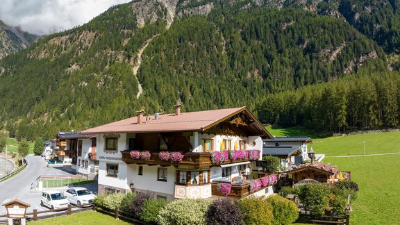 Ein charmantes Haus in den Bergen mit blühenden Pflanzen und einer schönen Aussicht auf die Natur. Im Hintergrund sind bewaldete Hügel und ein blauer Himmel zu sehen. - Apart Wiesengrund - Sölden