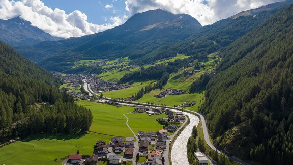 Eine malerische Berglandschaft mit sanften Hügeln und einem klaren Fluss. Im Vordergrund liegt ein kleines Dorf, umgeben von grünen Feldern und Wäldern. - Apart Wiesengrund - Sölden