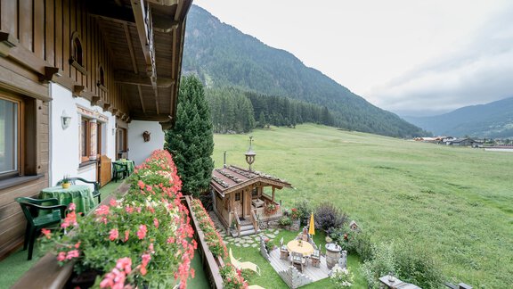 Eine malerische Ansicht von einem Balkon mit Blumen und Blick auf eine grüne Wiese und Berge im Hintergrund. Der Himmel ist bewölkt und es gibt einen kleinen Pavillon im Garten. - Apart Wiesengrund - Sölden