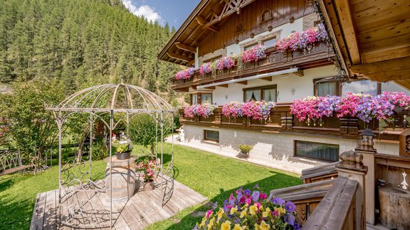 Ein charmantes Holzhaus mit Blumenbalkonen und einer schönen Gartenlandschaft. Im Vordergrund steht eine stilvolle Pergola umgeben von bunten Blumen. - Apart Wiesengrund - Sölden