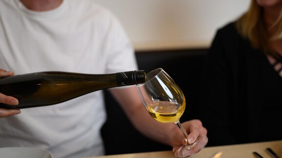Ein Mensch gießt Wein in ein Glas. Im Hintergrund ist eine andere Person zu sehen, die den Moment genießt. - Appartement Alpin - Sölden