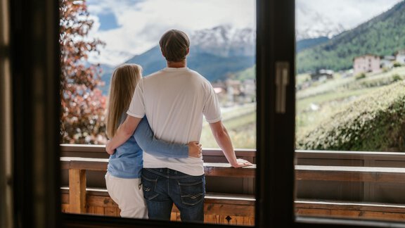 Ein Paar steht auf einem Balkon und blickt auf eine wunderschöne Berglandschaft. Im Hintergrund sind schneebedeckte Berge und grüne Wiesen zu sehen. - Appartement Alpin - Sölden