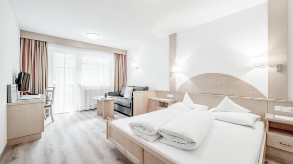 A modern room with a double bed, a television, and a cozy armchair. Large windows provide plenty of daylight and a pleasant atmosphere. - Appartementhaus Kathrin - Sölden