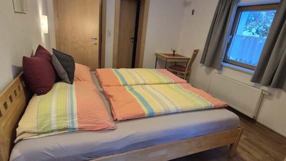Ein helles Schlafzimmer mit einem großen Bett und bunten Kissen. Im Raum befinden sich ein Tisch und ein Fenster mit Vorhängen. - Appt. Wildschütz - Sölden