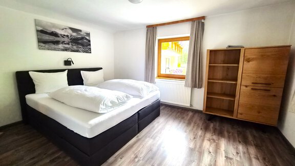 Ein helles Schlafzimmer mit einem Doppelbett und zwei Kissen. Im Raum steht ein Holzschrank und ein Fenster mit Aussicht nach draußen. - Arno`s Hoamat - Sölden