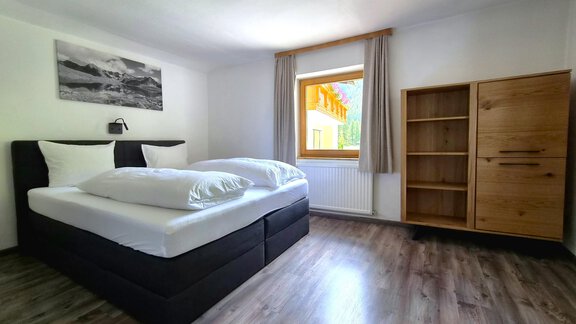 Ein helles Zimmer mit einem Doppelbett und zwei großen Kissen. Neben dem Fenster steht ein Holzregal und an der Wand hängt ein Bild. - Arno`s Hoamat - Sölden
