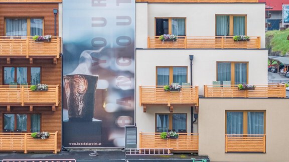 Ein modernes Gebäude mit einem großen Werbeplakat für ein Restaurant. Darunter befinden sich eine Terrasse mit Gästen und Fahrrädern. - Bäckelar Wirt - Sölden