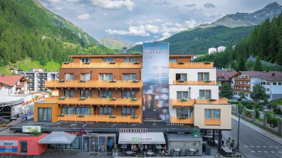 Ein modernes Hotelgebäude mit orangefarbenen Balkonen und einem großen Werbebanner. Im Hintergrund sind grüne Berge und ein blauer Himmel sichtbar. - Bäckelar Wirt - Sölden