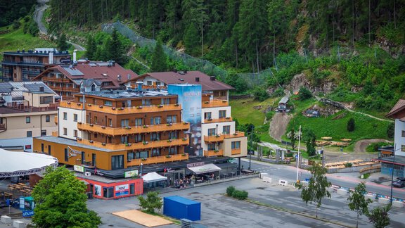 Eine malerische Berglandschaft mit modernen Gebäuden und einem Parkplatz. Umgeben von grünen Bäumen und sanften Hügeln. - Bäckelar Wirt - Sölden