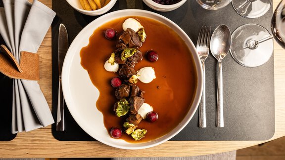 Ein schön angerichteter Teller mit Fleisch, Kräutern und verschiedenen Beilagen in einer dunklen Sauce. Im Hintergrund sind weitere Schalen mit Beilagen sichtbar. - Chalet Resort Sölden - Sölden