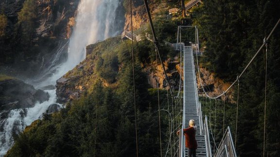 Eine Hängebrücke führt über eine Schlucht mit einem Wasserfall im Hintergrund. Grüne Bäume umgeben die Szene und verleihen ihr eine natürliche Schönheit. - Chalet White Pearl by MYALPS - Sölden