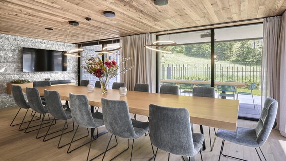 Ein moderner Konferenzraum mit einem großen Holztisch und grauen Stühlen. Große Fenster bieten einen Blick auf die grüne Umgebung und viel Tageslicht. - Chalet White Pearl by MYALPS - Sölden